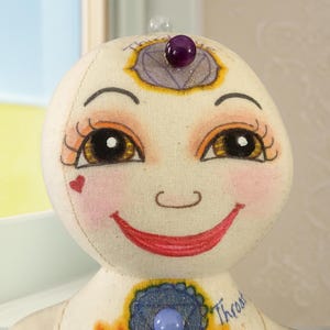 K&ouml;nnte beinhalten: Eine handgefertigte Puppe mit einem l&auml;chelnden Gesicht, bemalt mit warmen Farben. Die Puppe hat gro&szlig;e, ausdrucksstarke Augen, rosige Wangen und ein herzf&ouml;rmiges Detail. Verziert mit einem lila Edelstein auf der Stirn und Brust, verk&ouml;rpert sie ein spirituelles oder k&uuml;nstlerisches Thema.