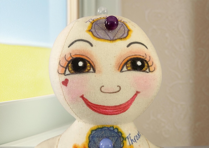 K&ouml;nnte beinhalten: Eine handgefertigte Puppe mit einem l&auml;chelnden Gesicht, bemalt mit warmen Farben. Die Puppe hat gro&szlig;e, ausdrucksstarke Augen, rosige Wangen und ein herzf&ouml;rmiges Detail. Verziert mit einem lila Edelstein auf der Stirn und Brust, verk&ouml;rpert sie ein spirituelles oder k&uuml;nstlerisches Thema.