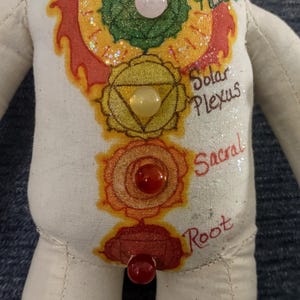 K&ouml;nnte beinhalten: Handgefertigte Puppe mit cremefarbenem K&ouml;rper, mit bemalten Chakra-Symbolen in leuchtenden Farben. Die Puppe hat blaue, rosa, gelbe und rote Edelsteinakzente. Die W&ouml;rter "Heart", "Solar Plexus", "Sacral" und "Root" sind auf der Puppe geschrieben.