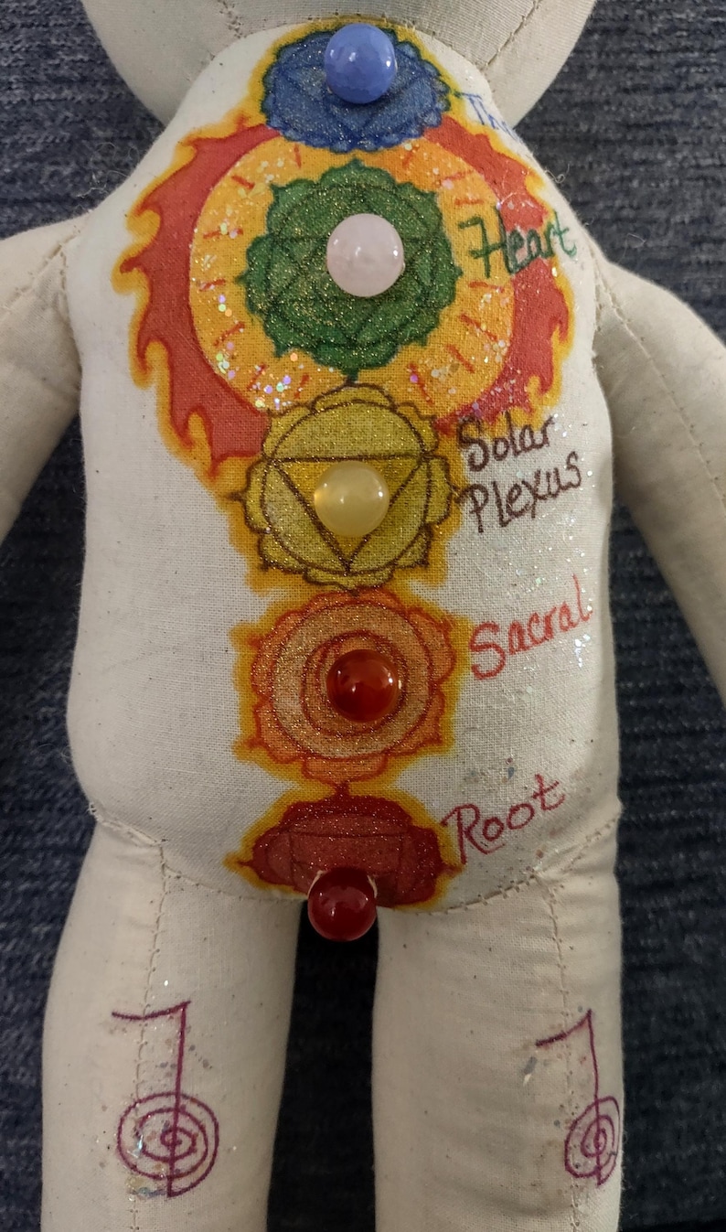 K&ouml;nnte beinhalten: Handgefertigte Puppe mit cremefarbenem K&ouml;rper, mit bemalten Chakra-Symbolen in leuchtenden Farben. Die Puppe hat blaue, rosa, gelbe und rote Edelsteinakzente. Die W&ouml;rter "Heart", "Solar Plexus", "Sacral" und "Root" sind auf der Puppe geschrieben.
