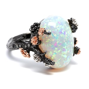 May include: A ring featuring a large, oval-shaped iridescent white opal cabochon set within a dark, textured metal band that resembles a tree branch, accented with small, raised rose gold and silver-toned floral or bud-like details.
