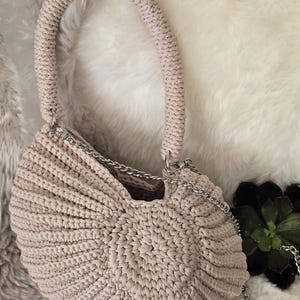 May include: A beige crochet handbag with a rounded, shell-like shape and a thick, textured handle. The bag features a central circular pattern with radiating ribbed stitches and includes a detachable silver-toned metal chain shoulder strap.