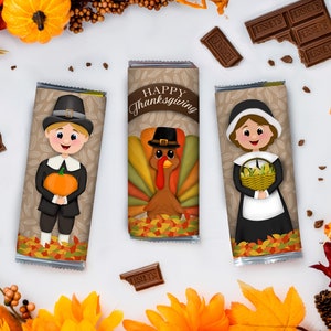 Könnte beinhalten: Drei Schokoladenriegel-Verpackungen mit Thanksgiving-Motiven. Die Verpackungen zeigen einen Pilgerjungen mit einem Kürbis, einen Truthahn und ein Pilgermädchen mit einem Korb Mais. Die Verpackungen sind mit Herbstblättern und dem Text "Frohes Thanksgiving" verziert.