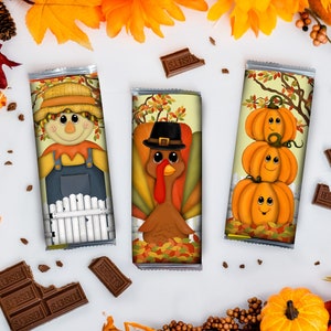 Op de afbeelding: Drie chocoladeverpakkingen met Thanksgiving-thema's. De verpakkingen tonen een vogelverschrikker, een kalkoen en een stapel pompoenen. De verpakkingen zijn op een witte achtergrond met herfstdecoraties.