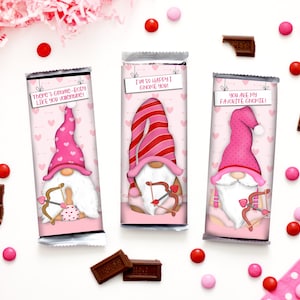 Puede incluir: Tres envoltorios de barritas de chocolate para el Día de San Valentín con gnomos con sombreros rosas y rojos y flechas en la mano. Los envoltorios tienen un fondo rosa con corazones blancos y el texto "There's gnome-body like you Valentine!", "I'm so happy I gnome you!" y "You are my favorite gnome!"