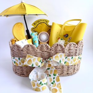 Pode incluir: Uma cesta trançada com borda festonada, envolta em uma fita branca com estampa de limões amarelos. A cesta contém um guarda-chuva amarelo, uma escova de cabelo amarela, uma sacola amarela, um pequeno ventilador portátil e um tubo com a etiqueta Brightening.