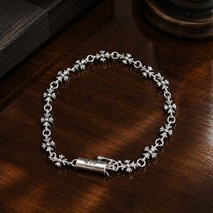 Könnte beinhalten: Ein silbernes Armband mit einer Reihe kleiner kreuzförmiger Anhänger. Das Armband ist durch kleine kreisförmige Glieder verbunden und hat einen Verschluss. Der Verschluss ist mit "925" graviert. Es liegt auf einer dunklen Holzoberfläche.