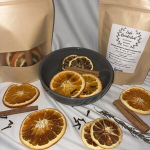 Peut inclure: Tranches d'orange séchées, bâtons de cannelle et clous de girofle sont disposés sur un tissu blanc, avec un bol gris contenant des éléments similaires. Deux sacs en papier brun remplis de tranches d'orange sont également présents. L'image est chaleureuse.