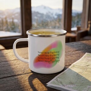 Könnte beinhalten: Ein weißer Emaillebecher mit silbernem Rand, gefüllt mit einer dampfenden Flüssigkeit. Der Becher zeigt einen abstrakten, aquarellartigen Hintergrund in Pink, Orange und Grün mit dem wiederholten schwarzen Text "Never Stop Exploring".