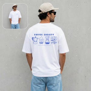 Op de afbeelding: Een wit t-shirt met korte mouwen en een blauwe afbeelding op de achterkant. Het ontwerp toont vier koffiezetapparaten met de tekst COFFEE SOCIETY en de zin GOOD COFFEE FOR A GOOD DAY in blauwe inkt op de stof gedrukt.