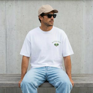 May include: A white short-sleeved t-shirt featuring a small graphic on the left chest with two green tennis rackets, a yellow tennis ball, and the text MATCH DAY in an arc above the rackets, paired with light blue denim jeans and a beige cap.