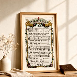 May include: A framed cross-stitch embroidery featuring the hymn Amazing Grace with musical notation, lyrics, and floral borders. The text includes the title Amazing Grace, verses, and the word Amen at the bottom, all centred within a light wood frame.
