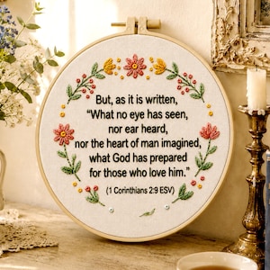 May include: A circular wooden embroidery hoop containing white fabric with black stitched text that reads, "But, as it is written, "What no eye has seen, nor ear heard, nor the heart of man imagined, what God has prepared for those who love him." (1 Corinthians 2:9 ESV)" surrounded by a floral wreath of pink, yellow, and green thread.
