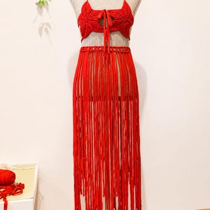May include: A bright red macrame two-piece set consisting of a halter-style crop top with star-shaped cups and a center tie closure, paired with a matching long skirt featuring a wide waistband and a floor-length fringe design made of vertical cords.