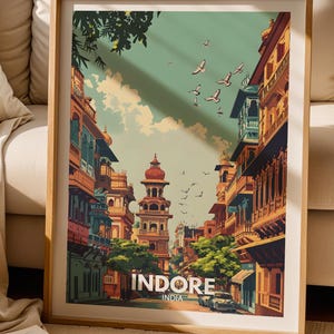 May include: A framed travel poster featuring an illustration of a street scene in Indore, India, with multi-storey buildings, green trees, and birds flying in a light sky. The text INDORE INDIA is printed in white at the bottom of the artwork.