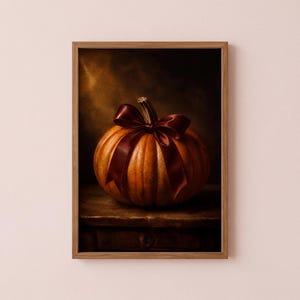 May include: A framed wall art print featuring a round orange pumpkin with a deep burgundy satin ribbon tied in a bow around its stem. The pumpkin rests on a dark wooden surface against a moody, shadowed background with warm golden lighting.