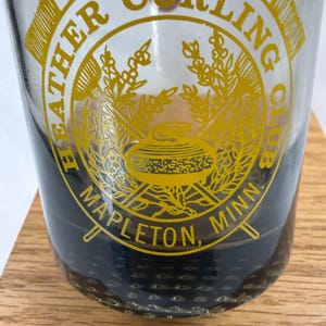 May include: A clear glass with a dark blue base and a gold logo. The logo reads "Heather Curling Club, Mapleton, Minn." with a curling stone graphic. The glass is sitting on a wooden surface.