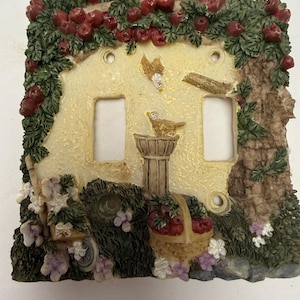 May include: A double light switch plate cover featuring a textured cream background with a raised relief design of an apple tree, a bird perched on a birdbath, a basket of red apples, and small purple and white flowers along the bottom edge.