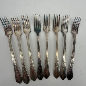 May include: A set of eight silver-toned metal forks arranged in a row. Each fork features four tines and a slender handle decorated with an embossed floral pattern near the base. The metal shows signs of natural tarnish and patina.