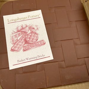 May include: A square, reddish-brown basket warmer with a woven brick pattern. A white label with the text "Longaberger Pottery" and "Basket Warming Bricks" is placed on the warmer. The warmer is inside a cardboard box.