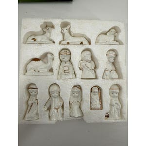 May include: A set of twelve white ceramic nativity figurines with gold-painted accents, including animals, angels, and figures, arranged in a white styrofoam storage tray. The pieces feature detailed facial expressions and clothing.