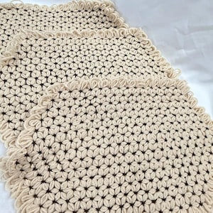 May include: Three cream-colored, square crocheted placemats. Each mat features a repeating floral pattern with dark brown centers and a fringed edge. The mats are arranged on a white surface.