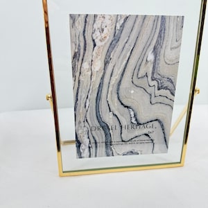 May include: A rectangular floating picture frame with a thin gold metal border and a clear glass pane. Inside, a display card features a gray and white marbled pattern with the text DREXEL HERITAGE and 5x7 printed in black ink.
