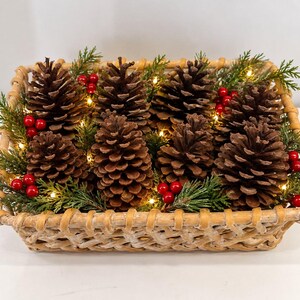 May include: A rectangular woven wicker basket filled with brown pinecones, green cedar sprigs, and clusters of bright red berries. The arrangement is illuminated by small, warm white string lights tucked between the natural elements.