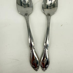 May include: Two silver-toned metal spoons featuring a polished finish and a decorative handle design. Each handle is engraved with a cursive letter B at the base, accompanied by subtle floral detailing near the tip of the spoon handle.