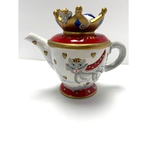 May include: A ceramic teapot featuring a white body with gold hearts, a gray cat illustration, and a red lid topped with a gold crown and a small mouse. The text "TIME FOR COFFEE" is printed in gold on the red lid. The base is red with gold hearts.