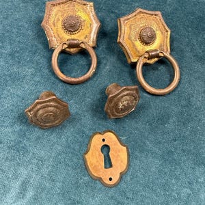 May include: Antique brass hardware set featuring two octagonal drawer pulls with ring handles, two smaller knobs, and a keyhole escutcheon. The hardware has a distressed, aged appearance, with a gold and brown color scheme.