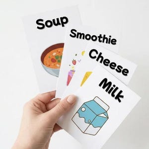 K&ouml;nnte beinhalten: Eine Hand h&auml;lt vier wei&szlig;e Karten mit Lebensmittelillustrationen und fettem schwarzem Text. Auf den Karten steht "Suppe", "Smoothie", "K&auml;se" und "Milch". Die Suppenkarte zeigt eine Sch&uuml;ssel mit Suppe. Die Smoothie-Karte hat ein Glas mit Fr&uuml;chten. Die K&auml;sekarte hat ein K&auml;seeck und die Milchkarte hat einen Milchkarton.