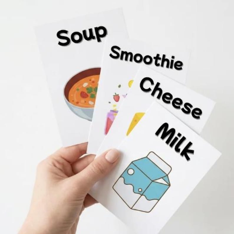 K&ouml;nnte beinhalten: Eine Hand h&auml;lt vier wei&szlig;e Karten mit Lebensmittelillustrationen und fettem schwarzem Text. Auf den Karten steht "Suppe", "Smoothie", "K&auml;se" und "Milch". Die Suppenkarte zeigt eine Sch&uuml;ssel mit Suppe. Die Smoothie-Karte hat ein Glas mit Fr&uuml;chten. Die K&auml;sekarte hat ein K&auml;seeck und die Milchkarte hat einen Milchkarton.