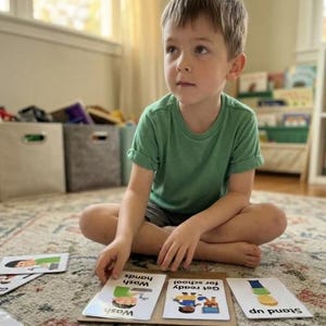 K&ouml;nnte beinhalten: Ein junges Kind sitzt auf einem gemusterten Teppich und interagiert mit Lernkarten. Die Karten mit Illustrationen und Text werden auf einer Kartonbasis angezeigt. Das Kind tr&auml;gt ein gr&uuml;nes T-Shirt und Shorts.