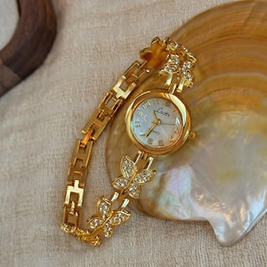 Könnte beinhalten: Eine goldfarbene Armbanduhr mit weißem Zifferblatt und goldenem Gliederarmband. Das Zifferblatt zeigt den Markennamen "Laotr" und das Wort "Quartz". Das Armband hat schmetterlingsförmige Akzente mit eingebetteten Kristallen.