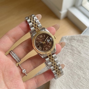 Könnte beinhalten: Eine zweifarbige Armbanduhr mit braunem Zifferblatt und goldfarbener Lünette und Akzenten. Die Uhr hat ein silbernes und goldenes Armband und trägt den Markennamen "BARSH" auf dem Zifferblatt. Die Uhr zeigt auch das Datum an.