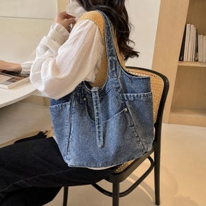 May include: A medium-wash blue denim tote bag featuring a structured silhouette, dual shoulder straps, and multiple exterior pockets with visible stitching details. The bag has a classic jean-like texture and a spacious main compartment opening.