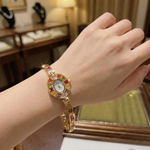 Puede incluir: Un reloj de pulsera dorado con una esfera circular y una correa adornada con gemas de colores. La esfera del reloj es blanca con delicadas manecillas y un pequeño logotipo. Las gemas son en tonos de rojo, amarillo, verde y morado.
