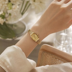 May include: A gold-toned wristwatch with a rectangular face and a linked band. The watch is worn on a wrist, showcasing its elegant design. The watch face features gold hands and markers, indicating the time. The watch band is approximately 6 inches in length.