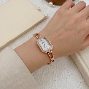 Puede incluir: Reloj de pulsera en tono oro rosa con esfera rectangular y bisel con cristales. El reloj tiene una esfera blanca con agujas doradas y la palabra "Luii" impresa. La correa del reloj también es de oro rosa y tiene un diseño de eslabones.
