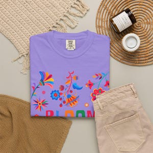 K&ouml;nnte beinhalten: Ein hellviolettes T-Shirt mit einem bunten Blumenmuster aus roten, blauen, orangen und gelben Bl&uuml;tenbl&auml;ttern. Das Wort BLOOM ist in gro&szlig;en, mehrfarbigen Buchstaben unten aufgedruckt. Das T-Shirt hat ein Comfort Colors Markenetikett am Kragen.