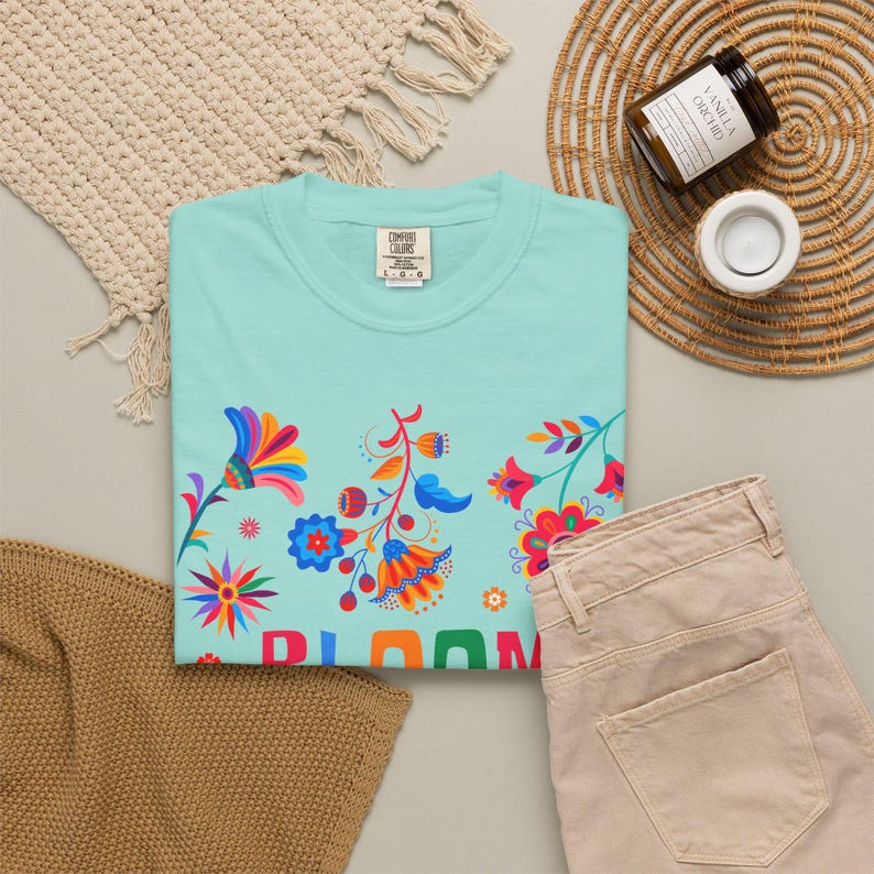 K&ouml;nnte beinhalten: Ein hellt&uuml;rkisfarbenes T-Shirt mit einem bunten Blumenmuster aus roten, orangen, blauen und rosafarbenen Bl&uuml;tenbl&auml;ttern und Bl&auml;ttern. Das Wort BLOOM ist in gro&szlig;en, bunten Blockbuchstaben am unteren Rand des Grafikdesigns auf der Vorderseite gedruckt.