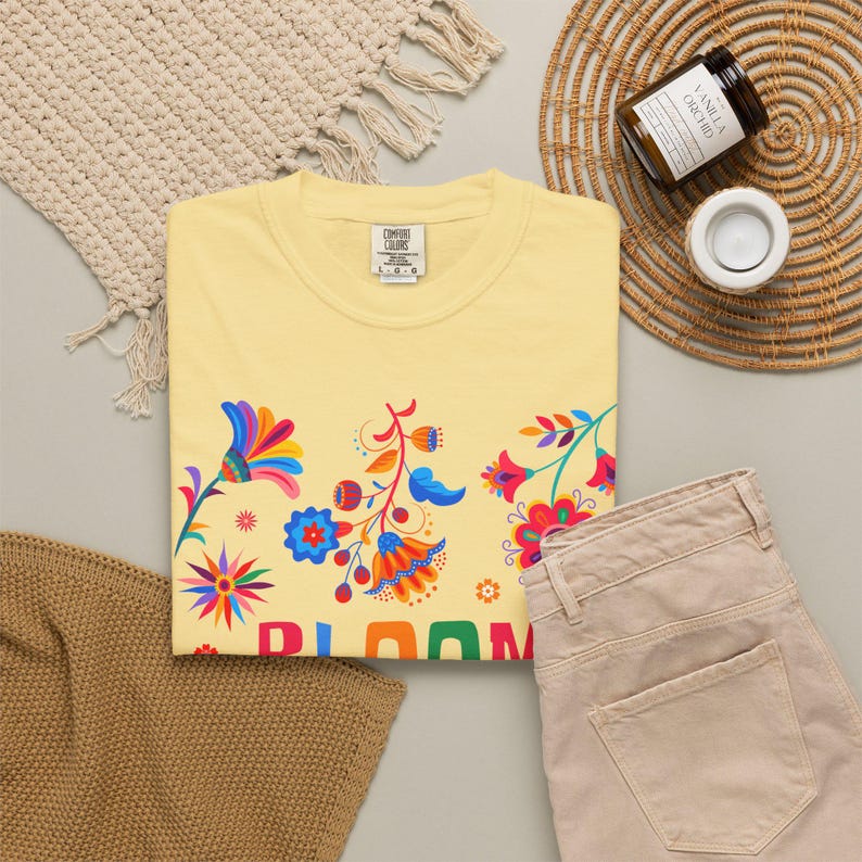 K&ouml;nnte beinhalten: Ein hellgelbes Kurzarm-T-Shirt mit einem bunten Blumenmuster aus roten, blauen, orangefarbenen und rosafarbenen Bl&uuml;tenbl&auml;ttern. Das T-Shirt hat ein Comfort Colors Markenetikett am Kragen und den Teiltext BLOOM in gro&szlig;en, bunten Buchstaben.