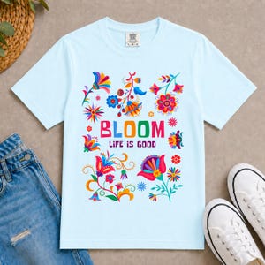K&ouml;nnte beinhalten: Ein hellblaues Kurzarm-T-Shirt mit einem bunten Blumenmuster und dem mittig aufgedruckten Text "BLOOM LIFE IS GOOD". Das Design zeigt verschiedene stilisierte Blumen in den Farben Rot, Pink, Gelb, Orange, Blau und Gr&uuml;n.