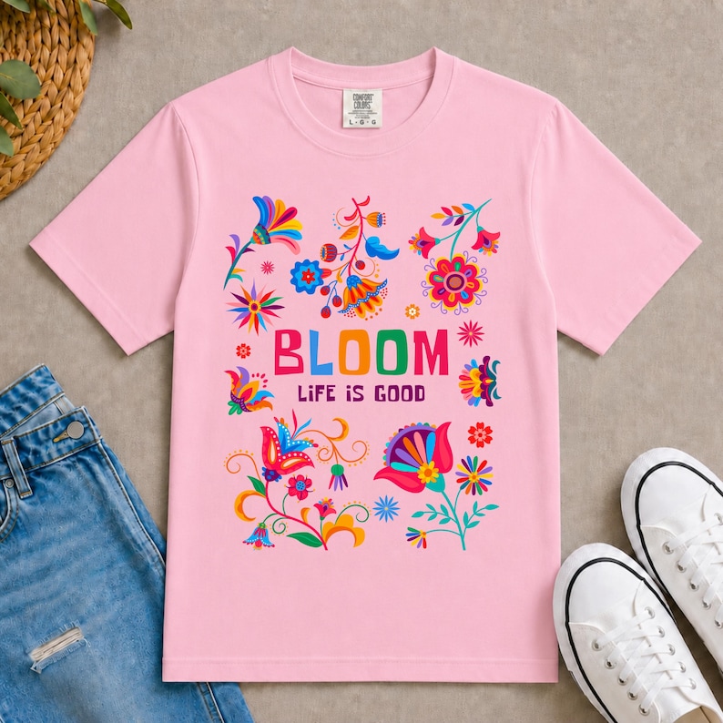K&ouml;nnte beinhalten: Ein hellrosa T-Shirt mit kurzen &Auml;rmeln, einem bunten Blumenmuster und dem mittig aufgedruckten Text "BLOOM LIFE IS GOOD". Das Design zeigt verschiedene stilisierte Blumen in den Farben Rot, Blau, Gelb, Orange und Violett.