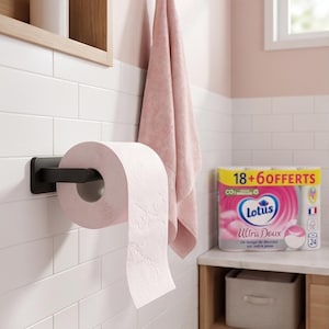 以下が含まれることがあります： 黒いホルダーにピンクのトイレットペーパーのロール、ピンクのタオル、棚にトイレットペーパーのパッケージがあるバスルームのシーン。壁はタイルの模様の白い壁です。