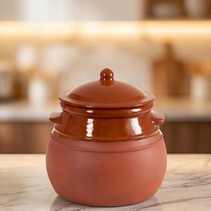 May include: A terracotta ceramic pot with a matching lid. The pot has a rounded body with two small handles and a darker band around the middle. The lid has a small knob on top. The pot is on a marble surface.