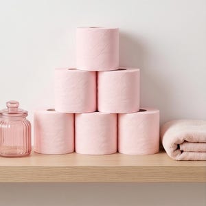 Puede incluir: Seis rollos de papel higi&eacute;nico rosa con un patr&oacute;n texturizado apilados en un estante de madera. Un peque&ntilde;o tarro de cristal rosa con tapa y una toalla rosa doblada tambi&eacute;n est&aacute;n en el estante, creando una est&eacute;tica de spa.