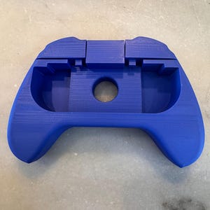 May include: A vibrant blue, 3D-printed gaming controller shell. The shell has a central circular opening and a textured surface. The design features a contoured shape for comfortable handling, with a focus on ergonomic design for extended use.