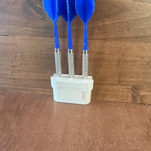 Op de afbeelding: Drie blauwe dartpijlen met zilveren metalen schachten worden getoond in een witte plastic houder. De darts zijn verticaal gerangschikt en tonen hun heldere blauwe kleur tegen een houten achtergrond. De houder heeft een rechthoekige vorm.